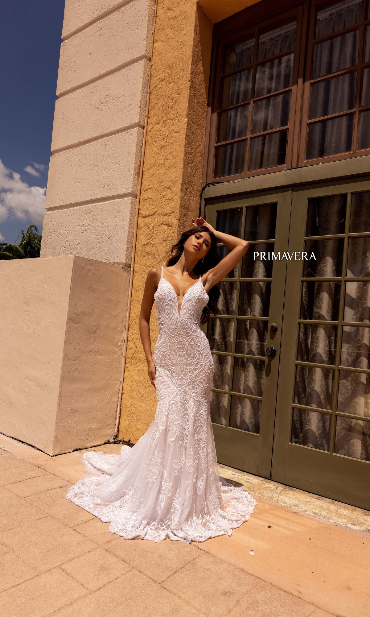 A woman poses outdoors in front of green double doors wearing the Long Wedding Dress 11105 by Primavera, an ivory mermaid gown with lace embroidery, intricate detailing, and a deep V neckline.